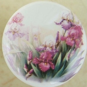 May include: A round, white ceramic lid featuring a floral design. The design showcases a cluster of purple and white iris flowers with green leaves. The flowers are painted in a watercolor style, with soft shading and delicate details.