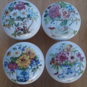 May include: Four white ceramic cabinet knobs with floral designs. Each knob features a different arrangement of flowers in a teacup, including roses, daisies, and other blooms. The knobs are round and have a glossy finish.