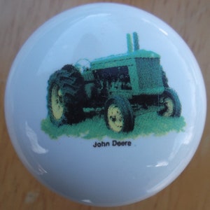 May include: A white ceramic cabinet knob featuring a green and black tractor design with the words "John Deere" printed below. The tractor is depicted on a green grassy area.