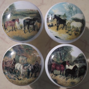 May include: Set of four white ceramic cabinet knobs. Each knob features a detailed, circular painting of horses in a pastoral landscape with trees and buildings. The horses are various colors, including brown, black, and white.