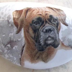 May include: White, oval-shaped ceramic object featuring a detailed illustration of a Boxer dog's face. The dog has brown fur and a black muzzle. A gray, abstract design is on the background. The object is likely a decorative item or a piece of art.