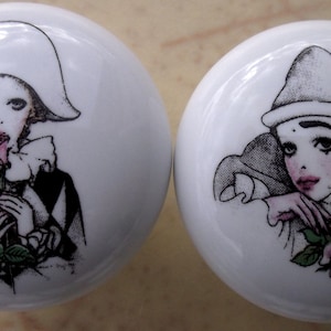 May include: Two white ceramic cabinet knobs with illustrations of Pierrot clowns. One clown wears a hat and diamond-patterned clothing, holding a pink rose. The other clown, also in a hat, rests their head on their hand, holding a pink rose.