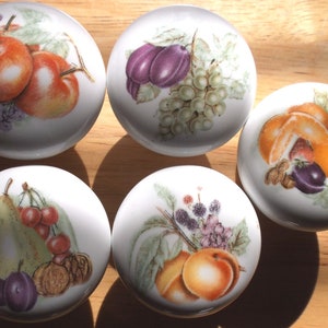 May include: Five white ceramic knobs with different fruit designs. Each knob features a different combination of fruits, including apples, pears, plums, grapes, oranges, and cherries. The knobs are perfect for adding a touch of whimsy to any kitchen or bathroom.