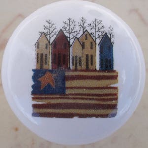 May include: A white ceramic knob with a colorful image of five houses and an American flag. The houses are red, yellow, and blue. The flag is red, white, and blue with a single star.