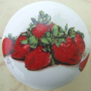 May include: A white ceramic knob with a colorful image of strawberries and green leaves. The strawberries are red and have a glossy finish.
