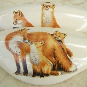 May include: Two white, oval-shaped decorative pieces with fox illustrations. One piece features a fox looking up, while the other depicts a family of foxes. The foxes are depicted in shades of orange, brown, and white.