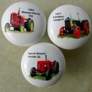 May include: Three white ceramic cabinet knobs with vintage tractor designs. One knob features a red 1954 Massey-Harris 745 tractor, another a 1917 Fordson model F, and the third a red David Brown model 25 tractor.