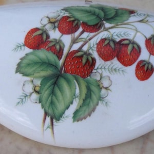 May include: White ceramic lid with a painted design of ripe red strawberries, green leaves, and white blossoms. The lid has a smooth, curved shape, and the strawberry design is centered on the surface. The overall theme is a kitchen or dining accessory.