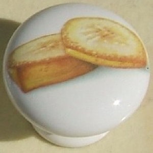 May include: A white ceramic knob with a painted image of two banana slices. The banana slices are yellow and brown.