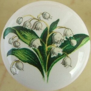 May include: White ceramic cabinet knob featuring a detailed illustration of lily of the valley flowers. The design includes white bell-shaped flowers, green leaves, and stems. The knob is round and has a glossy finish.