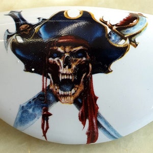 May include: A white ceramic door knob with a colorful pirate skull and crossbones design. The skull is wearing a blue and gold pirate hat with a red bandana.