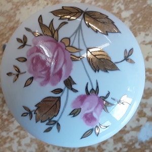 May include: A white ceramic cabinet knob with a floral design. The knob features pink roses and gold-colored leaves and accents. The design is set against a white background, creating a classic and elegant look. The knob is round.