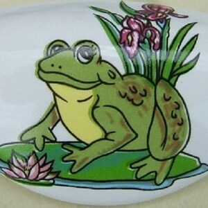 May include: A white ceramic knob with a green frog sitting on a lily pad. The frog has brown spots and is surrounded by green leaves and pink flowers.