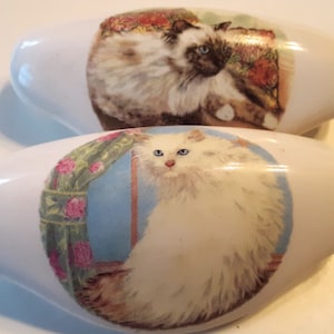 May include: Two white ceramic cabinet knobs with oval shapes. Each knob features a detailed illustration of a fluffy cat. One cat is brown and white, the other is white. The knobs are likely for furniture or decorative use.