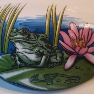 May include: A white, oval-shaped decorative item featuring a green frog on a lily pad, a pink lotus flower, and reeds against a blue water background. The design is painted in a cartoon style.