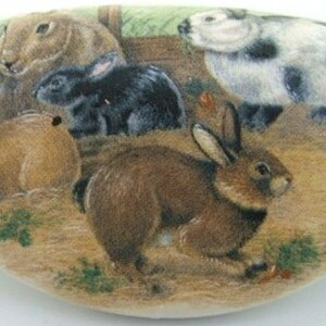 May include: Hand-painted ceramic tile featuring a whimsical scene of four rabbits in a grassy field. The rabbits are brown, black, white, and brown with black spots. The tile is oval-shaped and has a white background.
