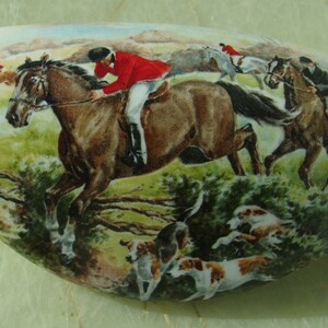 May include: A white ceramic oval-shaped dish with a painted scene of a horse race. The scene features three riders on horseback, two hounds, and a green landscape.