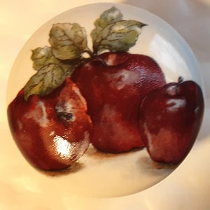 May include: A hand-painted ceramic tile featuring three red apples with green leaves. The apples are painted in a realistic style with highlights and shadows.