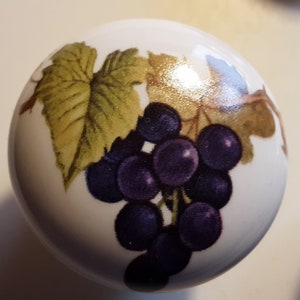 May include: A white, round decorative knob featuring a cluster of purple grapes and green and gold leaves. The design is printed on the surface of the knob, which appears to be ceramic or porcelain.