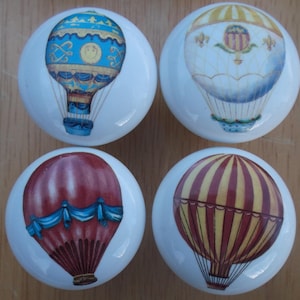 May include: Four white ceramic cabinet knobs with hot air balloon designs. The balloons are in shades of blue, red, yellow, and gold, with intricate details. Each knob has a rounded shape, perfect for adding a decorative touch to furniture.