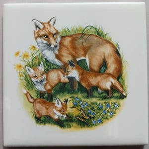May include: A square ceramic tile featuring a family of foxes in a grassy field with yellow flowers and blue flowers. The adult fox has reddish-brown fur and is surrounded by three fox kits. A butterfly is also present.