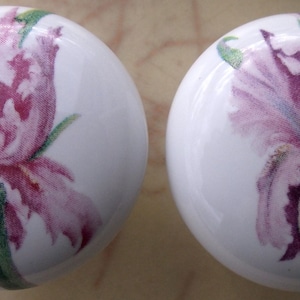 May include: Two white ceramic knobs with a pink and purple iris flower design. The flowers are painted in a watercolor style.