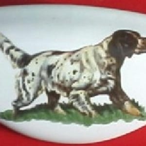 May include: White, oblong decorative item with a painted image of a dog. The dog is a brown and white setter breed, walking on a patch of green grass. The background is white, and the item is set against a red surface.