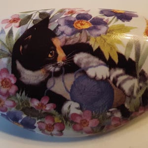 May include: White ceramic soap dish with a painted design of a black and white cat playing with a blue ball of yarn, surrounded by pink and purple flowers and a butterfly. The dish has an oval shape.