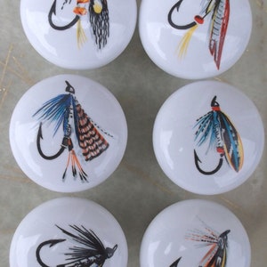 May include: Six white ceramic knobs with colorful fishing fly designs. Each knob features a different fly pattern with a hook.