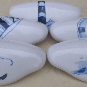 May include: Five white ceramic drawer pulls with blue nautical designs. The designs include an anchor, a lighthouse, a sailboat, a life preserver, and a ship's wheel.