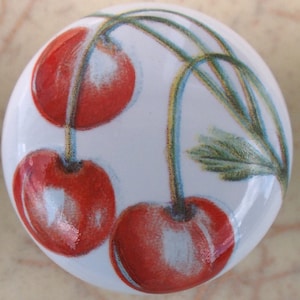 May include: Ceramic knob with a white background and a design of three red cherries with green stems and leaves.