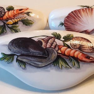 May include: Three white, oval-shaped decorative stones. Each stone features a hand-painted design of seashells, shrimp, and greenery. The shells are in shades of brown, and the shrimp are orange. The stones are arranged on a white surface.