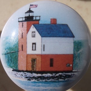 May include: A round, white ceramic knob featuring a detailed illustration of a lighthouse. The lighthouse has a red brick tower, a white house section, and a blue roof. An American flag waves atop the tower, with water and sky in the background.
