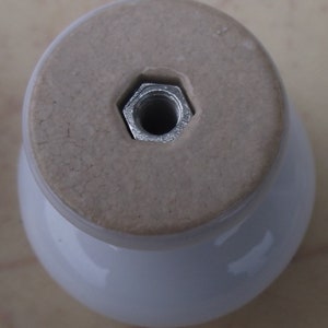 May include: A white ceramic cabinet knob with a rounded base and a flat, light tan top. A silver, hexagonal nut is embedded in the top for attachment. The knob has a glossy finish.