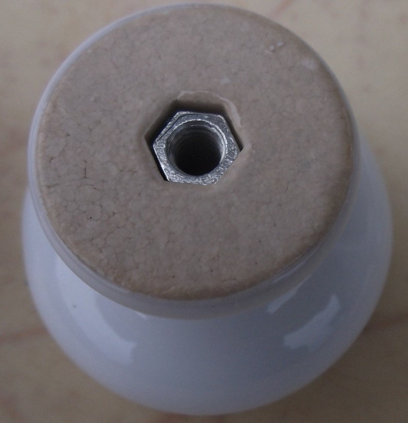 May include: A white ceramic cabinet knob with a rounded base and a flat, light tan top. A silver, hexagonal nut is embedded in the top for attachment. The knob has a glossy finish.
