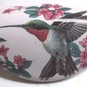 May include: White ceramic decorative piece with a hummingbird and pink flower design. The hummingbird has green, red, and gray feathers, with its wings spread. Pink flowers and green leaves are painted around the edges of the piece.