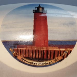 May include: A white, oval-shaped decorative item featuring a red lighthouse with a black top, set against a blue sky and water. The base of the lighthouse is on a wooden pier. The text "Milwaukee Pierhead, WI" is displayed on a banner.