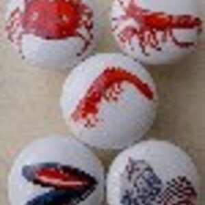 May include: Five white golf balls with various sea creature designs. The designs include a red crab, a red lobster, a red shrimp, a black and white mussel, and a blue and white fish. The golf balls are arranged in a circular pattern.