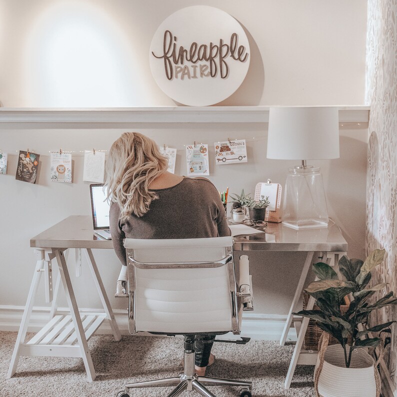 May include: A woman sits at a silver desk in a home office, working on a laptop. A white sign with the words "fineapple pair" hangs on the wall. The office is decorated with plants, artwork, and a white lamp.