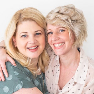 May include: Two women with blonde hair are smiling and hugging. They are both wearing polka dot shirts. The woman on the left is wearing a green shirt and the woman on the right is wearing a white shirt.