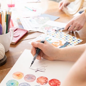 May include: A person is using a brush pen to create watercolor art on white paper. The table also holds art supplies, including paintbrushes, colored pencils, and a spool of twine. Colorful watercolor designs are visible.