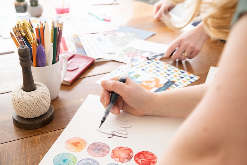May include: A person is using a brush pen to create watercolor art on white paper. The table also holds art supplies, including paintbrushes, colored pencils, and a spool of twine. Colorful watercolor designs are visible.