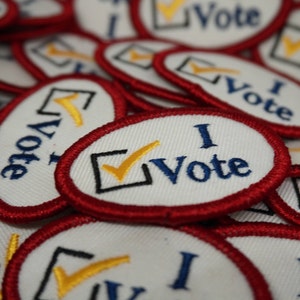 Op de afbeelding: Close-up van een stapel witte stoffen patches met rode randen. Elke patch heeft een zwart vinkje met een gele vinkje erin en de tekst "I Vote" in blauwe borduurwerk.