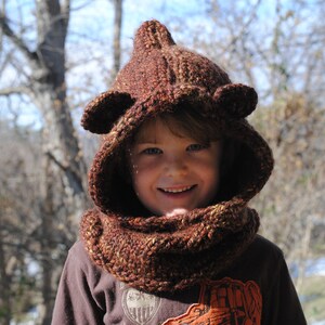 May include: A brown knitted hooded scarf with ear flaps. The hood is pulled up over the head of a child, and the scarf is pulled up around their neck. The child is wearing a brown shirt.