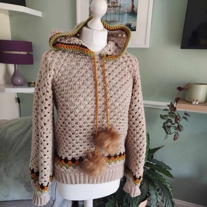 May include: A beige crocheted hooded sweater with brown, orange, and green trim. The sweater has a drawstring hood and two brown pom-poms on the front.