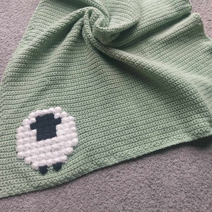 May include: A green crocheted baby blanket with a white sheep applique. The sheep has black eyes and a black nose.