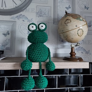 Puede incluir: Una rana de crochet verde con ojos blancos sentada en un estante de madera. La rana tiene patas largas y pies grandes. El estante está sobre una superficie de azulejos negros.