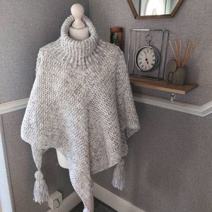 Pode incluir: Um poncho de malha branco e cinza com gola alta e franjas. O poncho é exibido em um manequim.