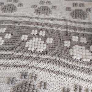 May include: A crocheted blanket with a repeating pattern of paw prints. The blanket is made with white and gray yarn and has a textured, puffy look.