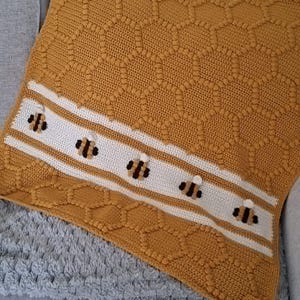 May include: A mustard yellow crochet blanket featuring a textured honeycomb stitch pattern throughout. A horizontal white stripe runs across the blanket, decorated with five small, three-dimensional crocheted bees with black and yellow striped bodies.